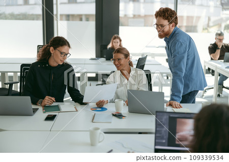 Group of young business people discussing something and smiling while sitting at the office table Group of young business people discussing something and smiling while sitting at the office table 109339534