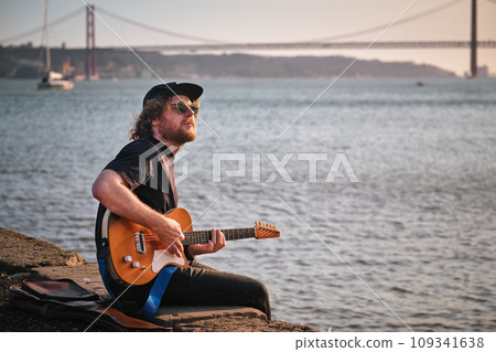 Hipster street musician in black playing electric guitar in the street sitting on pier embankment on sunset Hipster street musician in black playing electric guitar in the street sitting on pier embankment on sunset 109341638