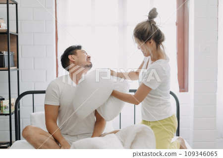 Funny couple angry wife hit husband with pillow in white bedroom. Funny couple angry wife hit husband with pillow in white bedroom. 109346065