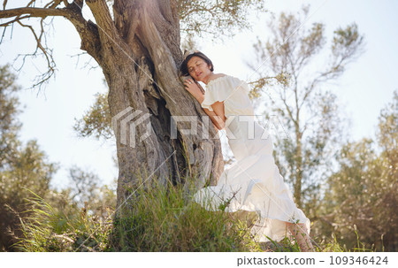 Beautiful Asian young woman in white dress outdoor near olive tree. embracing fresh air and engaging in outdoor activities. Friluftsliv concept means spending as much time outdoors as possible 109346424