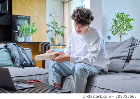 Young male unpacking an online purchase, sitting on couch at home Young male unpacking an online purchase, sitting on couch at home 109353663