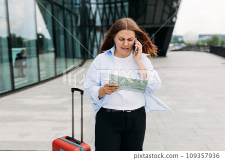 Unhappy 30s woman tourist dressed in casual look speaking on smartphone gadget on the city street. Angry redhead girl with a red suitcase and map standing on city street. Negative people emotion. 109357936