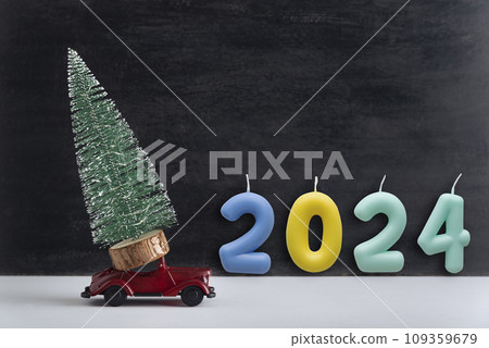 Toy car carries miniature Christmas tree and colored numbers 2024, New Year. Delivery of Christmas trees concept 109359679