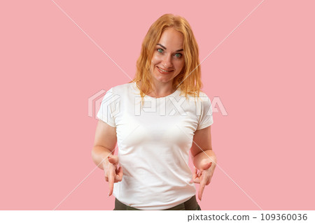 Smiling woman points her fingers down on pink background looks in camera. Portrait of young smiling round-faced woman with ginger hair. 109360036