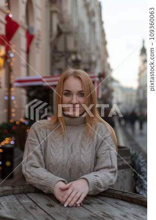 Attractive woman in sweater on the street cafe. Christmas decorations in the background. Cold season, walk in the city. Vertical frame 109360063