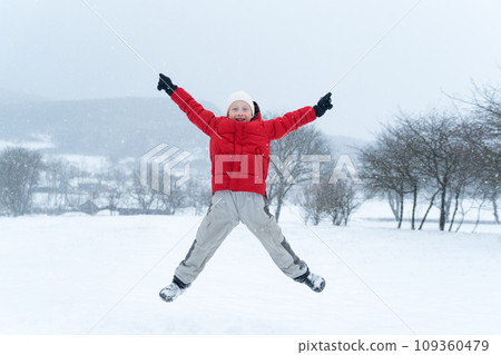 Child with his hands up and enjoys the winter holidays. Teenage boy in red jacket jumps during snowfall Child with his hands up and enjoys the winter holidays. Teenage boy in red jacket jumps during snowfall 109360479