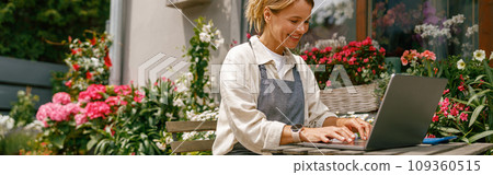 Small flower shop owner florist wearing apron working on laptop and taking online orders in store Small flower shop owner florist wearing apron working on laptop and taking online orders in store 109360515