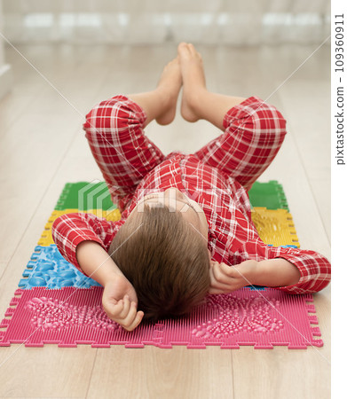 Sport and health concept. A little boy 4 years old in red checkered pajamas is working out on a multi-colored massage orthopedic mat with spikes in a home interior. Close-up. Sport and health concept. A little boy 4 years old in red checkered pajamas is working out on a multi-colored massage orthopedic mat with spikes in a home interior. Close-up. 109360911