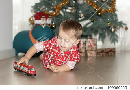 Christmas concept. New Year 2024. A small handsome boy in red checkered pajamas lies on the floor and plays with a red steam locomotive against the background of a decorated Christmas tree with gifts. 109360935