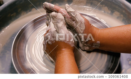Soiled hands of a female potter working with a piece of white clay on a potter's wheel. Pottery workshop setting. The concept of manual labour, art and craftsmanship. Soiled hands of a female potter working with a piece of white clay on a potter's wheel. Pottery workshop setting. The concept of manual labour, art and craftsmanship. 109361408