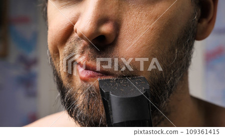 A man trims his beard with a trimmer. Close up. An electric razor trimming a beard. Black beard with grey hairs. Beard care at home. A man trims his beard with a trimmer. Close up. An electric razor trimming a beard. Black beard with grey hairs. Beard care at home. 109361415