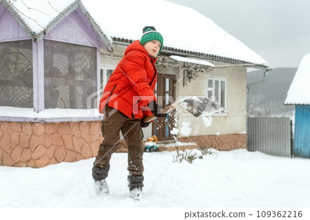 Little boy in warm puffy jacket and knitted hat with shovel cleans the snow in the yard of house. Snowy winter, cold weather Little boy in warm puffy jacket and knitted hat with shovel cleans the snow in the yard of house. Snowy winter, cold weather 109362216