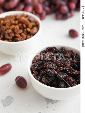 Bowls of brown and dark raisins with ripe red grapes on light background. Bowls of brown and dark raisins with ripe red grapes on light background. 109362719