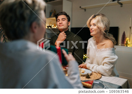 Rear view of unrecognizable middle-aged woman receiving Christmas gift from loving family at home on xmas eve. Loving daughter giving present to mother on holidays. Concept of festive atmosphere. Rear view of unrecognizable middle-aged woman receiving Christmas gift from loving family at home on xmas eve. Loving daughter giving present to mother on holidays. Concept of festive atmosphere. 109363546