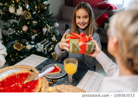 Back view of loving grandmother giving festive box with Christmas present to adorable granddaughter sitting at dinner feast table during xmas family party. Concept of home festive atmosphere. 109363553