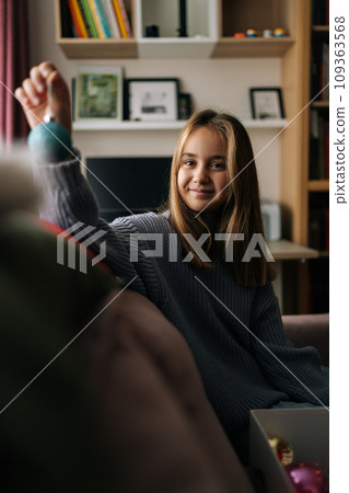 Vertical portrait of adorable little girl preparing for holidays, choosing festive Christmas tree toys from box, decorates xmas tree for New Year in living room, smiling looking at camera. 109363568