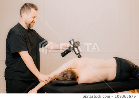 A therapist massages a man back with a massage percussion device in her home. The therapist's hand holds a therapeutic vibrating massager. Physical therapy and muscle recovery and massage 109363679