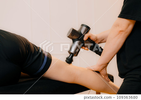 A therapist massages a man back with a massage percussion device in her home. The therapist's hand holds a therapeutic vibrating massager. Physical therapy and muscle recovery and massage 109363680