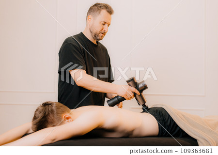 A therapist massages a man back with a massage percussion device in her home. The therapist's hand holds a therapeutic vibrating massager. Physical therapy and muscle recovery and massage 109363681