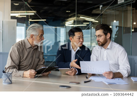 Three diverse business people talking together sitting at a meeting desk inside office, businessmen in business suits discussing with boss business owner about financial indicators, achievements. Three diverse business people talking together sitting at a meeting desk inside office, businessmen in business suits discussing with boss business owner about financial indicators, achievements. 109364442