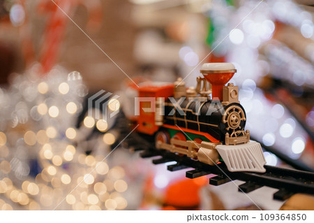 toy vintage steam locomotive on the floor under a decorated Christmas tree on a background of bokeh lights garland. Family fun, joy, activity for little kids. Children's new year holiday. toy vintage steam locomotive on the floor under a decorated Christmas tree on a background of bokeh lights garland. Family fun, joy, activity for little kids. Children's new year holiday. 109364850