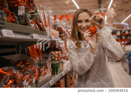Cute young woman choosing artificial Christmas tree in Xmas store. Christmas concept store, buying ornaments. A woman buys New Year's decorations for a Christmas tree in a store. Cute young woman choosing artificial Christmas tree in Xmas store. Christmas concept store, buying ornaments. A woman buys New Year's decorations for a Christmas tree in a store. 109364858