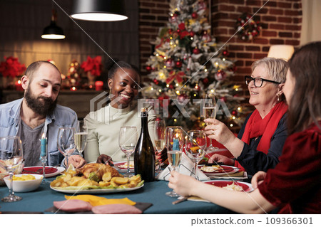 Grandma makes toast at dinner table with friends and family, enjoying christmas eve festivity at home. Senior woman giving speech with raised wine glasses for cheers, winter celebration. 109366301