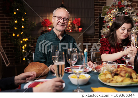 Grandpa at dinner table with family eating traditional meal and celebrating christmas holiday at home. Senior adults meeting together at festive gathering during winter seasonal event. Grandpa at dinner table with family eating traditional meal and celebrating christmas holiday at home. Senior adults meeting together at festive gathering during winter seasonal event. 109366344