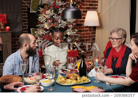People raising glasses for toast during christmas dinner, enjoying family time at home with traditional food. Diverse friends making speech to thank everyone for celebrating around the table. People raising glasses for toast during christmas dinner, enjoying family time at home with traditional food. Diverse friends making speech to thank everyone for celebrating around the table. 109366357