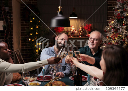 Diverse persons clinking alcohol glasses at christmas eve dinner, celebrating holiday tradition with wine and homemade meal. Friends and family saying cheers with drinks at table, cozy ornaments. 109366377