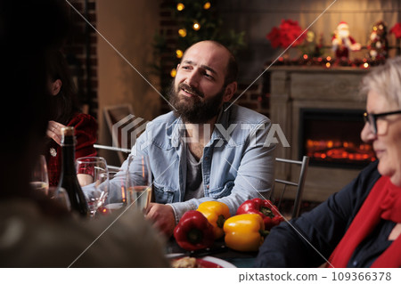 Happy persons talking about life and enjoying december holiday at home, grandparents and family members having fun eating traditional food and drinking wine. Diverse people gathering around table. 109366378