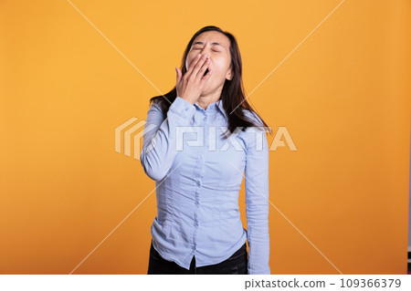 Portrait of sleepy filipino woman covering mouth with hand being tired while posing in studio standing over yellow background. Exhausted model yawning having lazy expression, working overtime 109366379