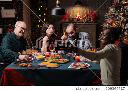 Persons taking photos at festive dinner, making memories of christmas eve celebration at home. Diverse friends and family having fun with drinks and food, taking pictures with smartphone. Persons taking photos at festive dinner, making memories of christmas eve celebration at home. Diverse friends and family having fun with drinks and food, taking pictures with smartphone. 109366387