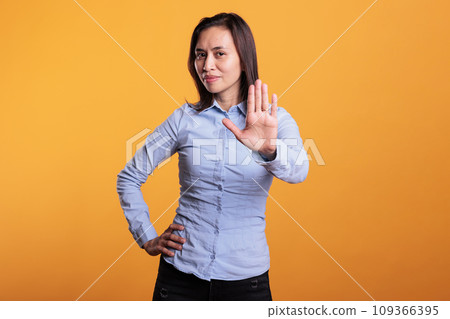 Serious filipino woman doing rejection gesture with hand in front of camera, posing in studio over yellow background. Negative model showing stop gesture, being in denial and expressing refusal. 109366395
