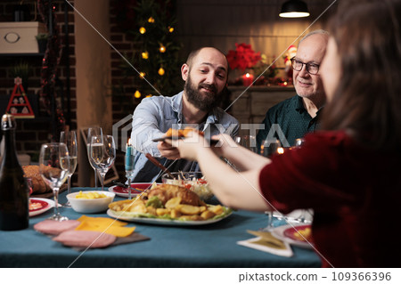 Persons eating food at festive dinner, celebrating christmas eve together and passing meal plates around. Joyful group of people enjoying holiday celebration event at home, winter feast. Persons eating food at festive dinner, celebrating christmas eve together and passing meal plates around. Joyful group of people enjoying holiday celebration event at home, winter feast. 109366396