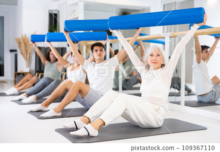 Men and women practicing pilates with roller on gray mat in gym room Men and women practicing pilates with roller on gray mat in gym room 109367110