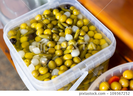 Pickled olives, onion and cucumbers in plastic container on market. Popular vegetarian appetizer closeup 109367252