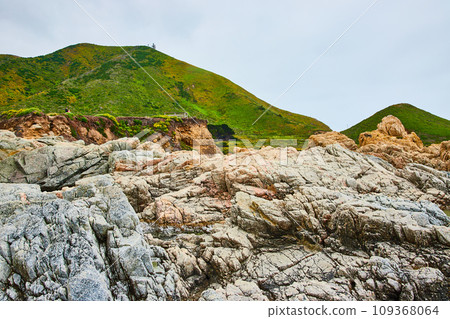 Baren rock cliff face in jagged crags with distant green mountainous hillside and overcast sky 109368064