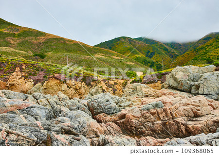 Baren rock cliff face with jagged crags and distant green mountainous hills under overcast sky 109368065