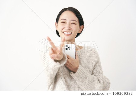 Middle-aged woman holding a smartphone and making a peace sign, white background 109369382