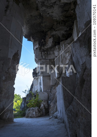 A rectangular opening in a dark, mossy limestone cave frames a bright blue sky with fluffy white clouds in Setigi, Gresik, East Java, Indonesia. 109370141
