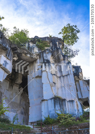 A weathered limestone cliff, adorned with a smattering of trees, stands tall against a clear blue sky in Setigi, Gresik, East Java, Indonesia. 109370530
