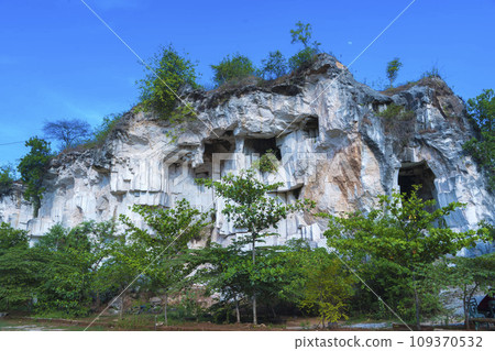 A weathered limestone cliff, adorned with a smattering of trees, stands tall against a clear blue sky in Setigi, Gresik, East Java, Indonesia. 109370532
