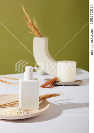 An unlabeled bottle of shower gel is placed on a ceramic plate with whole grain rice. Decorations are displayed on a white table with a moss green background. Mockup for new product advertising. 109372056