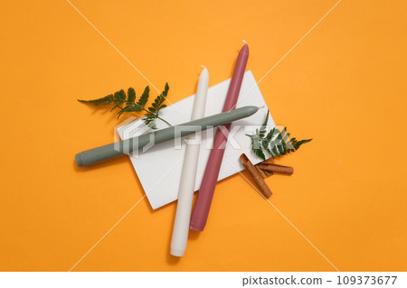 Colored candles and green leaves are placed on a white podium on a yellow background. Creative space for advertising. Product advertising with minimalist background. 109373677