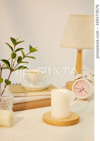Candle is placed on a round wooden platform. Green leaves are placed in a glass vase. Coffee cups, notebooks, lamp and clock are displayed against a minimalist background. Copy space. 109373678