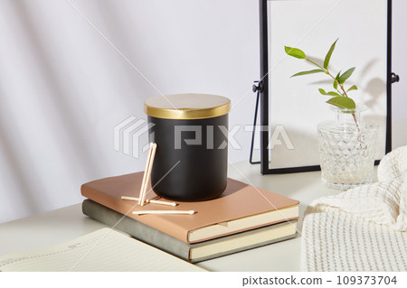 A jar of unlabeled scented candles is placed on top of two notebooks. A mirror, glass flower vase and cloth towels are displayed on the table. Delicate white background for scented candle advertising. 109373704