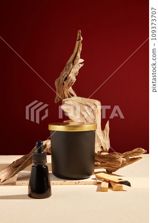 A bottle of unlabeled scented candles is placed in the center of the frame, surrounded by dry branches and a bottle of black serum. Beige and red background. Mockup for scented candle advertising. 109373707