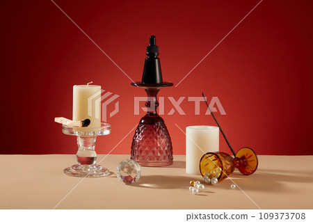 A black serum bottle sits on glass candlesticks amid decorative cups, white candles, and pearls on a beige table. Set against a red backdrop, a creatively inviting space for messaging. 109373708