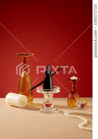 A black serum bottle is placed on a glass candlesticks, decorative glass cups, white candles and a string of pearls are displayed on a beige table. Red background. Creative space for copying. 109373710